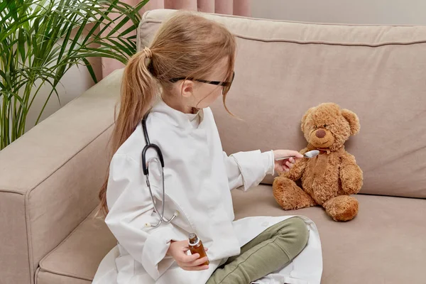 Una chica rubia juega la profesión de médico y le da un juguete a un osito de peluche . —  Fotos de Stock