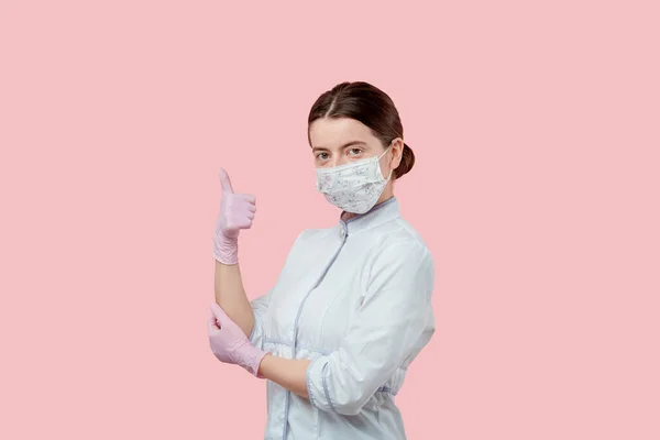 Μελαχρινή κοπέλα δείχνει σούπερ χειρονομία, αντίχειρες επάνω σε ένα ιατρικό παλτό, γάντια και μάσκα. — Φωτογραφία Αρχείου