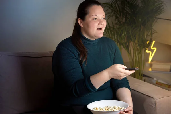 Ung vacker flicka tittar på TV på natten och äter popcorn. Kroppsligt positiv. — Stockfoto