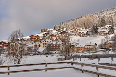 Ön planda kar içinde ahşap bir çit var. Karla kaplı renkli evlere bak.