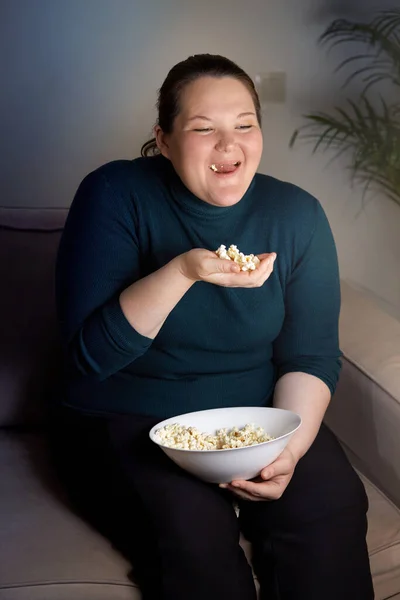 Vertikalt foto av en flicka som skrattar, äter popcorn och tittar på TV. — Stockfoto