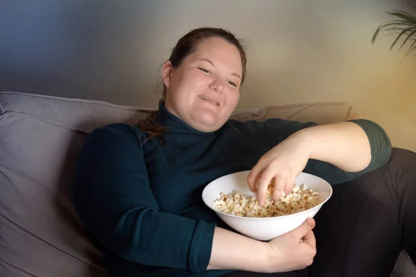 Een lachend meisje met overgewicht in de woonkamer kijkt tv met popcorn.. — Stockfoto