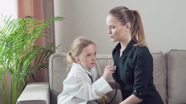 Una niña interpreta a un médico y escucha la respiración de la madre con un fonendoscopio . — Vídeos de Stock