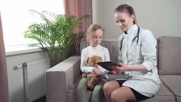 Une femme médecin est assise sur un canapé et montre une fille de dessin animé sur une tablette. — Video