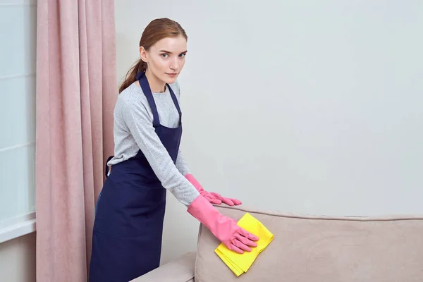 Limpeza de casa profissional. Menina olha para a câmera e lava o sofá com pano . — Fotografia de Stock