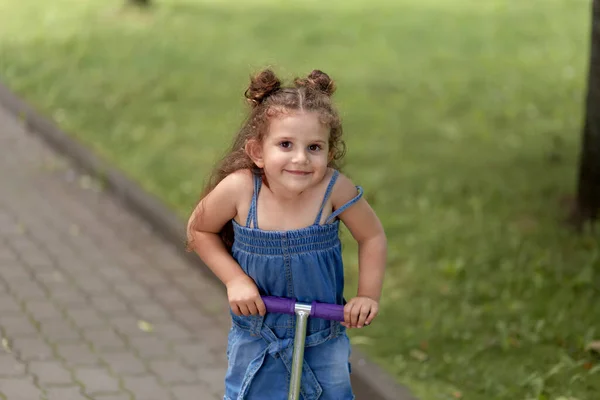 Menina feliz com cabelo ondulado monta uma scooter em um dia ensolarado no parque . — Fotografia de Stock