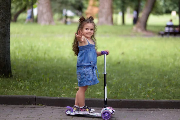 Menina feliz com cabelo ondulado monta uma scooter e acena a mão. Uma menina amigável . — Fotografia de Stock