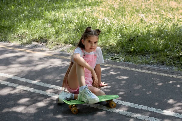 Menina bonita com longos cabelos lisos escuros senta-se na pista com um skate . — Fotografia de Stock