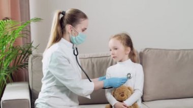 Bir kız kanepeye oturur ve elinde bir oyuncak tutar. Çocuk doktoru tarafından muayene edilir..