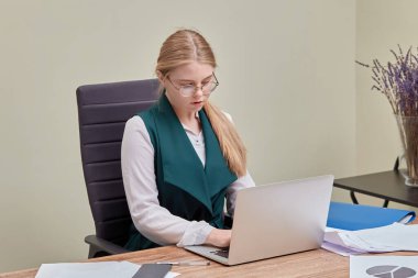 Şirin sarışın bir kız ofiste bir sandalyede oturup dizüstü bilgisayara bakıyor..