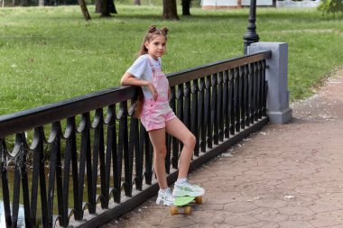 Uzun saçlı güzel bir kız çitin yanındaki kaykayın üstünde duruyor..