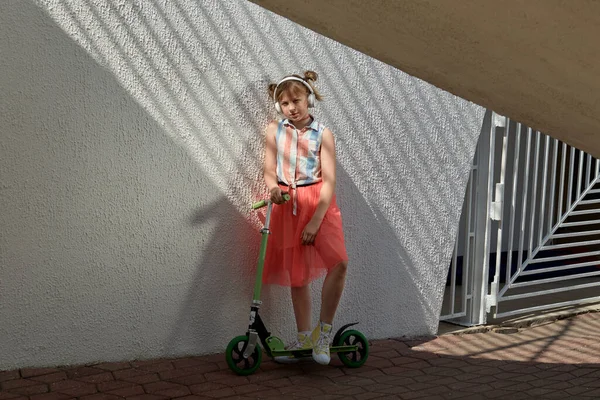 Uma garota bonita ouve música e monta uma scooter ao sol. Um dia ensolarado brilhante . — Fotografia de Stock