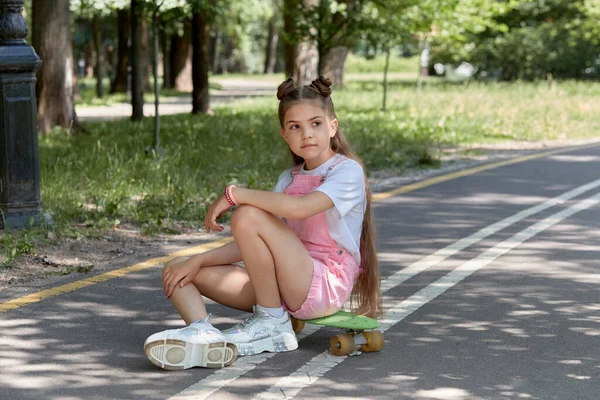 Uma linda garota bonita está sentada na pista com um skate em suas mãos . — Fotografia de Stock