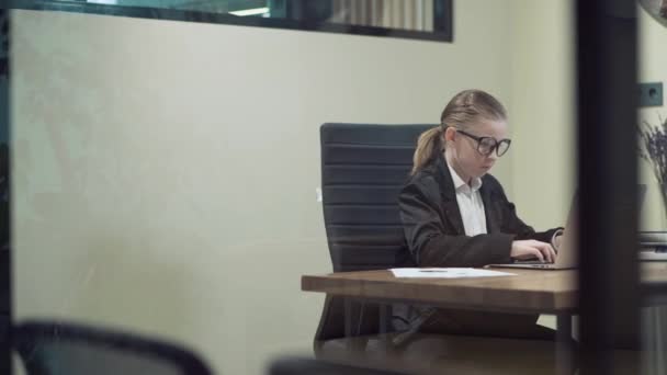 За склом, мила дівчина в окулярах працює в офісі і дивиться на її щоденник . — стокове відео