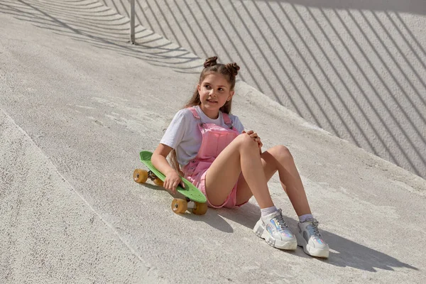 Uma menina bonita brilhante em um macacão na moda com um skate olha para o lado — Fotografia de Stock