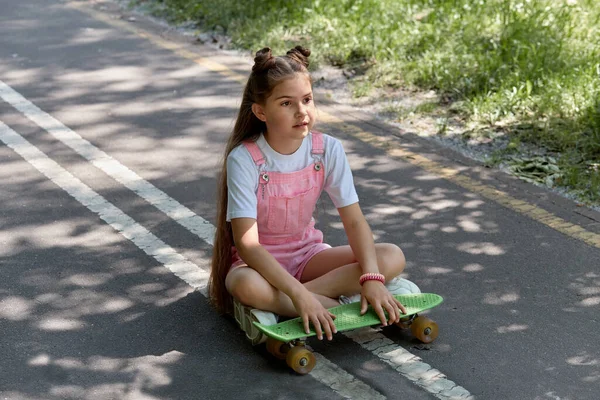 Uma linda garota bonita está sentada na pista com um skate em suas mãos . — Fotografia de Stock