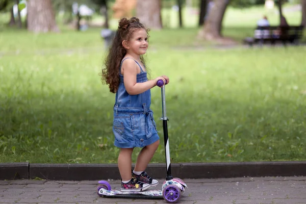 Menina feliz com cabelo ondulado monta uma scooter em um dia ensolarado no parque . — Fotografia de Stock