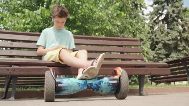 Un chico guapo está sentado en un banco, sus piernas están en un scooter giratorio . — Vídeos de Stock