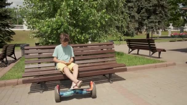 A handsome guy sits on a bench in a summer park, his feet are on a gyroscooter. — Stock Video