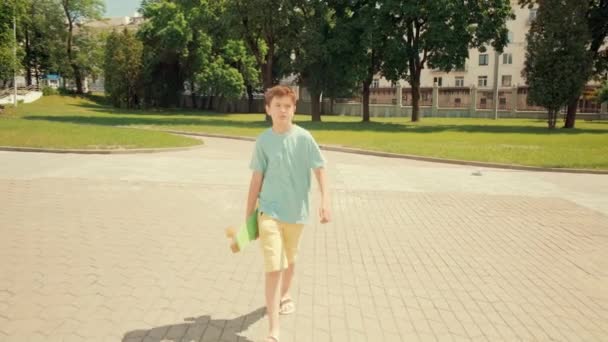 한 갈색의 소년이 스케이트보드를 손에 들고 길을 따라 걷고 있다. 태양이 내리는 날씨. — 비디오