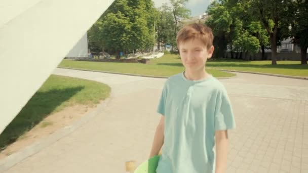 행복하게 잘 생긴 갈색 머리 소년이 스케이트보드를 손에 들고 걷고 있습니다. 태양이 내리는 날씨. — 비디오