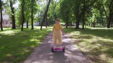 Güzel, mutlu bir kız zarif bir elbiseyle parkta scooter sürüyor..