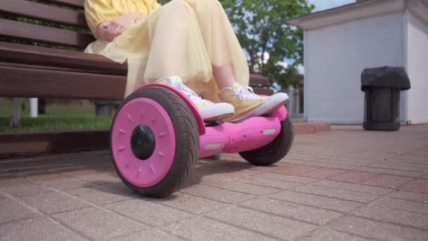 Beine eines Mädchens, das auf einer Bank sitzt, Beine auf einem Hoverboard. Sommerpark. — Stockvideo