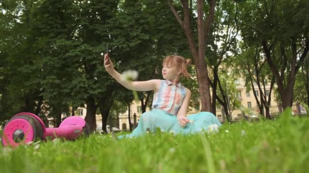 Una chica con auriculares escucha música, se sienta en la hierba y se toma una selfie . — Vídeos de Stock