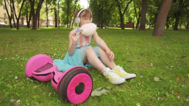 Girl in headphones listens to music, sits on the grass next to a gyro scooter. — Stock Video