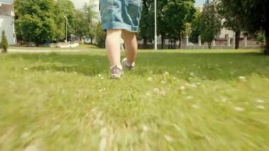 Küçük bir kızın bacakları parlak yeşil çimlerde spor ayakkabılarla yürüyor. Yaz tatili