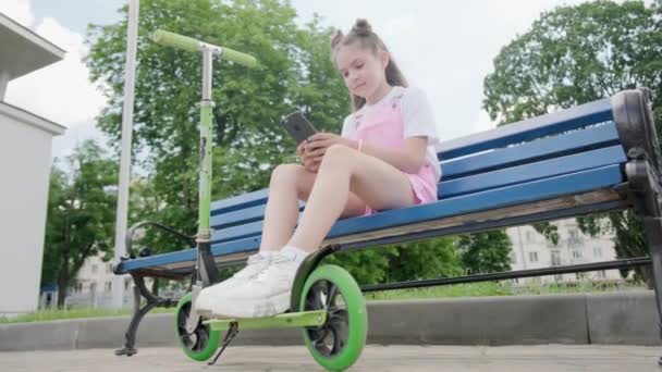 Vista inferior de una chica sentada en un banco en el parque con un teléfono en las manos . — Vídeos de Stock