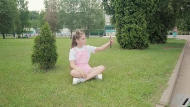 더운 여름날 풀밭에 앉아 있는 귀여운 소녀가 셀카를 만든다. — 비디오