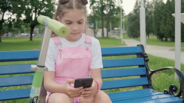 Uma menina se senta em um banco com um telefone e se comunica nas redes sociais . — Vídeo de Stock