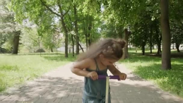 Een meisje rijdt vooruit op een kick scooter in een zomerpark. pluizen en stofvliegen. — Stockvideo