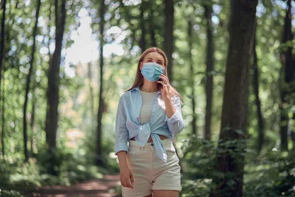 Uma menina caminha no parque usando uma máscara e fala em um smartphone móvel . — Fotografia de Stock