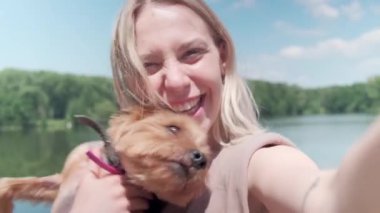 Sarışın kız köpeğini tutuyor ve gölün arka planında fotoğraflarını çekiyor..