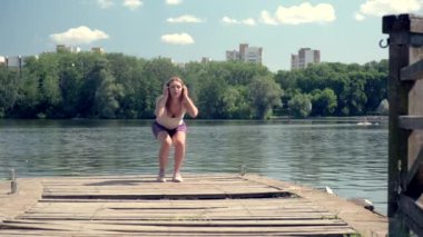 Çömelme hareketleri. Kızın atletik bir vücudu var. Sporcu göl kenarındaki iskelede..