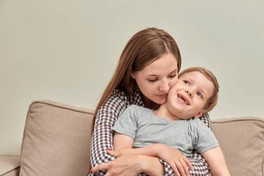 Genç, güzel esmer anne oğluna sarılıyor ve yanağından öpüyor. Gülümseyen çocuk