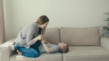 Genç güzel anne ve bebek kanepede eğlenip oynuyorlar. İç mekan. 4k