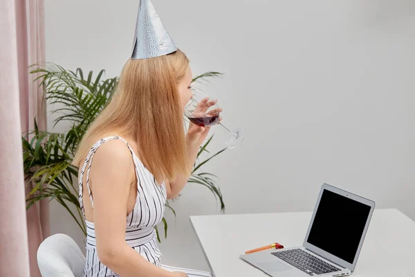 Flicka med en festlig mössa på huvudet framför laptop med ett glas rött vin. — Stockfoto