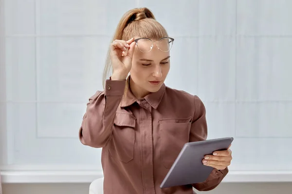 Uma jovem mulher em óculos para a vista olha atentamente para o tablet . — Fotografia de Stock