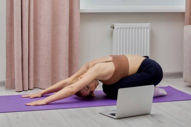 Genç bir kadın laptopun yanında yoga ve spor yapıyor..