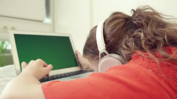 Una chica cansada en los auriculares se encuentra boca abajo en la cama mira la pantalla del ordenador portátil. — Vídeo de stock