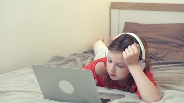 Uma linda garota deitada na cama está estudando online com fones de ouvido brancos. — Vídeo de Stock