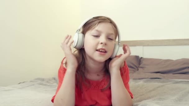 女の子はヘッドフォンで音楽を聞いてベッドの上に横たわっています。オンラインコミュニケーション. — ストック動画