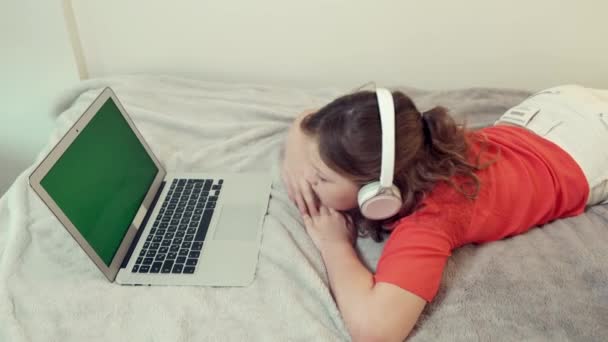 美しいです女の子はベッドの上に横たわっていますオンラインで勉強しています白いヘッドフォン. — ストック動画