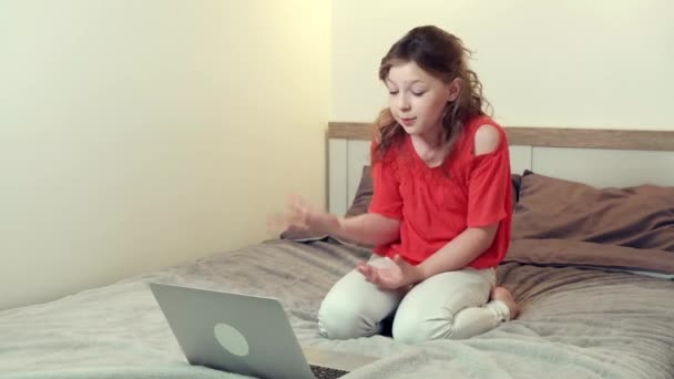 Egy aranyos lány fekszik az ágyon, előtte egy laptop, egy videó link segítségével. — Stock videók