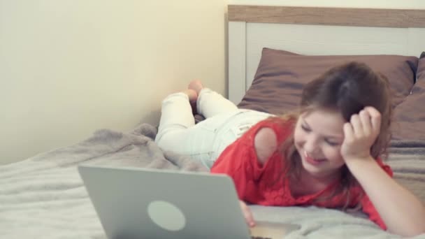 幸せな女の子はベッドの上にあり、ビデオ通話で友人と話します. — ストック動画