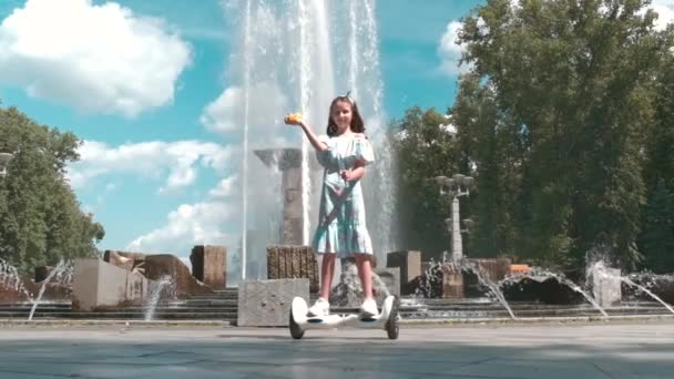 Ένα κορίτσι σε ένα φόρεμα βόλτες με ένα hoverboard και φυσάει φυσαλίδες στο πάρκο το καλοκαίρι. — Αρχείο Βίντεο