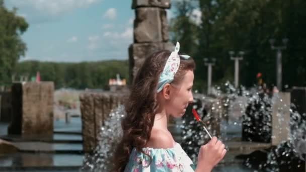 Una chica con un vestido largo cerca de la fuente. Hay un caramelo en un palo. — Vídeo de stock